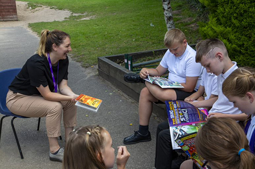 A group of children sat outside looking at books and magazines with their teacher who is smiling.