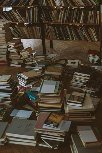 Photograph of a bookcase wth books also piled onto the floor.