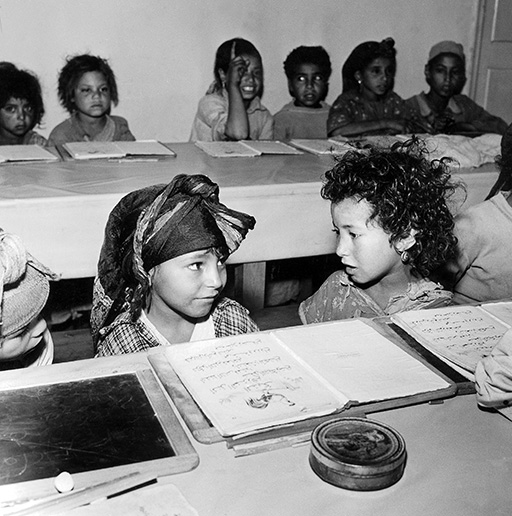 A black-and-white photograph of a group of children learning in a school setting.