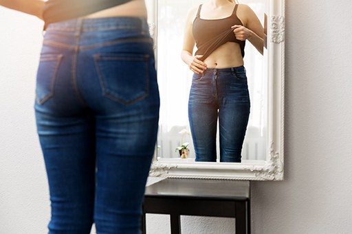 An image of a woman exposing her abdomen and looking in a full length mirror.
