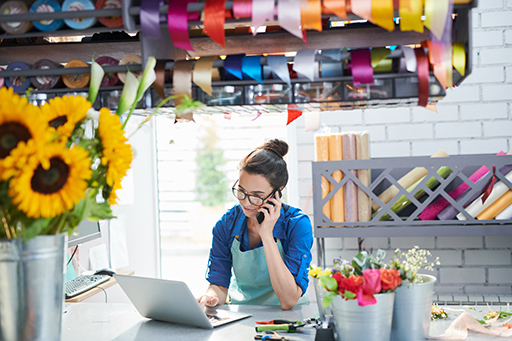 Women on the phone looking at her laptop in a room containing crafts and flowers.
