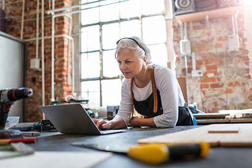 A woman leaning over a workbench looking at a laptop.