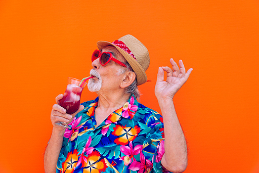 Grey-haired man wearing a floral Howaiian-style shirt, heart-shaped sunglasses, sipping a cocktail.
