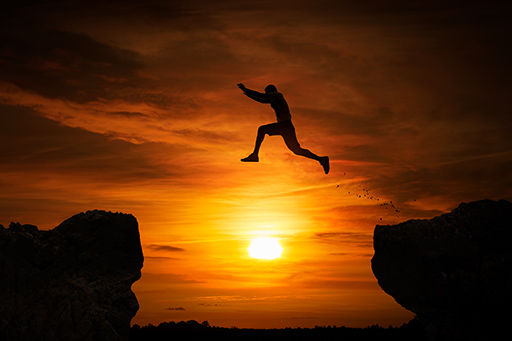 A silhouette of a person leaping from one rock to another with the sun setting in the background.