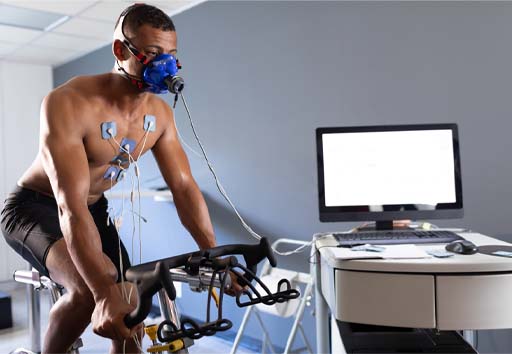 The image shows a man on a cycle ergometer with electrodes on his chest and a mask on his face. He is completing a fitness test.