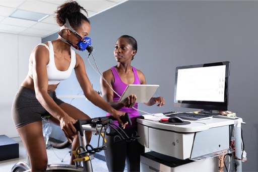 The image shows a woman on a cycle ergometer with electrodes on her chest and a mask on her face. She is completing a fitness test and being shown results by a female tester.