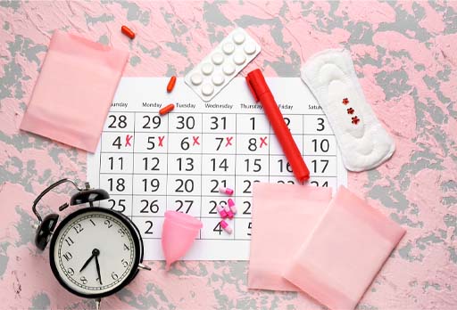 The image shows products related to the menstruation – sanitary towels and pads, pills, a calendar and pen, an alarm clock and a cap.