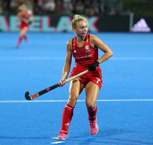 The image shows a female hockey player. She is holding a hockey stick across her body and is ready to receive the hockey ball.