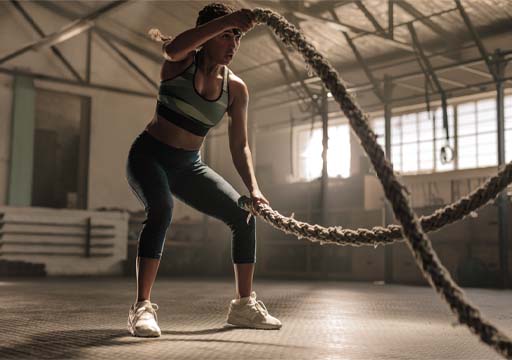 The image shows an athletic female working out in a gym. She is completing a battling ropes exercise.