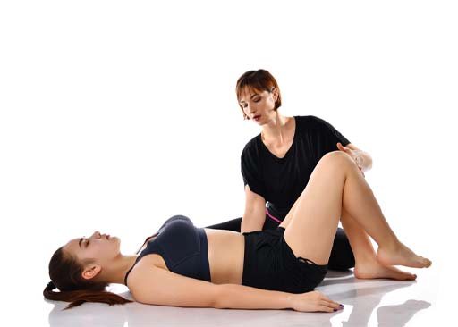 The image shows two females, one of which is lying on her back with her knees bent and other one has one hand behind the back of the female lying down and the other one her knee. She is teaching the female how to train her pelvic floor muscles.