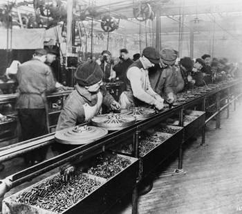 A production line of workers is shown in a black and white photograph from 1913. The workers all wear hats and are arranged in a line behind a conveyor belt that's got wheel like pieces on it and boxes below that appear to be full of screws.