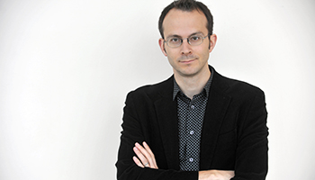 Photograph of Tim Harford looking head on at the camera through his glasses. He's wearing a black suit and a black and white tightly dotted shirt.