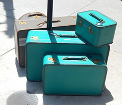 Three turquoise and two grey suitcases stacked together on the pavement. 