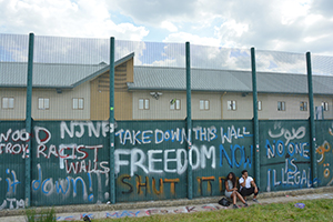 Image of the wall outside Yarl's Wood with protest graffiti all over it, and two young people sit outside. 
