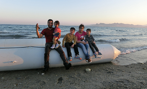A family sit on the edge of a boat on the beach, smiling - having arrived on the Greek island of Kos. 