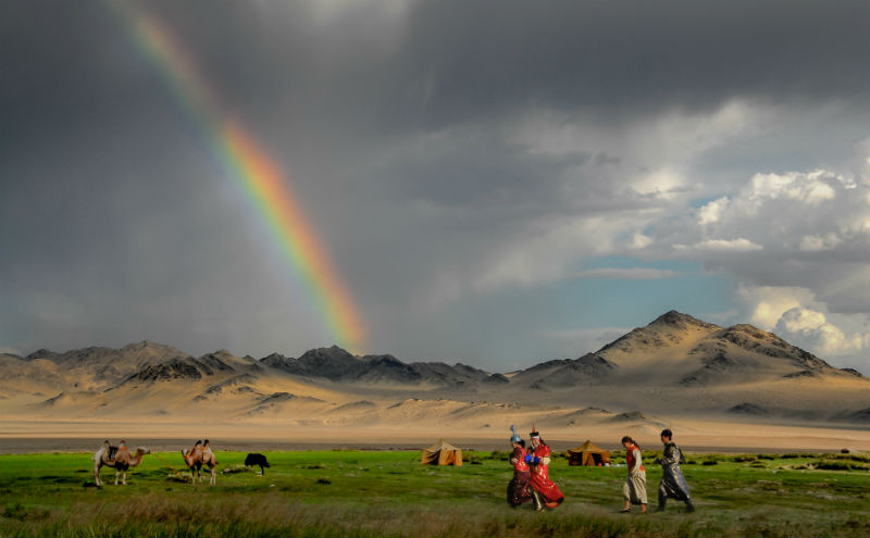 Monglian people dressed for the Naadam Festival cross in front of a rainbow