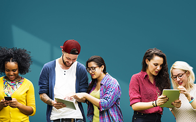 five young people in vibrant clothes look at tablets and phones together in front of a blue background. 