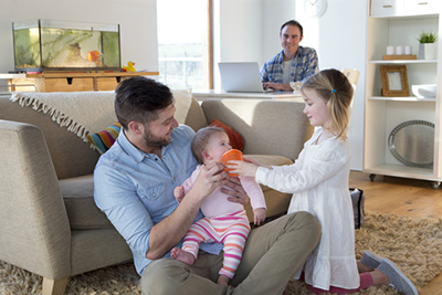 Same sex male couple at home with their two daughters. One father is sitting on the floor with their baby girl in his arms. The older daughter is helping with the baby. The other father is sitting at a table in the background with a laptop in front of him and is watching his family.