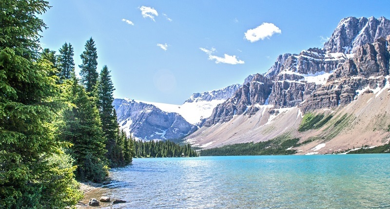 Photograph of Moraine lake, Alberta. The photo shows a blue-green expanse of lake water, with dense forest on the left, and rocky mountainous terrain to the right of the lake.