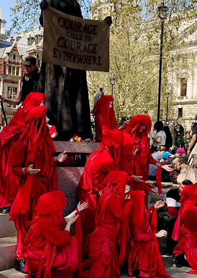 Nine people are dressed in red to symbolise blood through costume. They are wearing hoods and the four at the front are kneeling down whilst the other five are stood on the steps of a statue. There are people in the background.