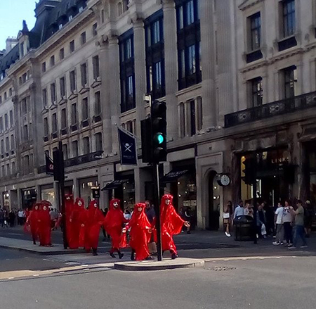 Nine people are dressed in red to symbolise blood through costume. They are wearing hoods and the four at the front are kneeling down whilst the other five are stood on the steps of a statue. There are people in the background.