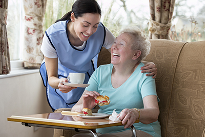 A young female carer bringing a cup of tea to a smiley elderly lady