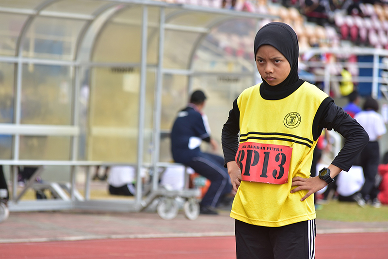 A young child stands at the beginning of a track with hands on her hips looking ahead determinedly. She has a yellow track bib on and 'BP13' is marked in black on a background of red. 