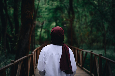 The image shows back of a person wearing a hijab looking at trees as she walks on through them. She is wearing a white t-shirt and a black satchel over one shoulder. She is walking into a woodland area with dark trees.