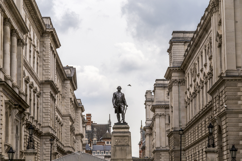 A statue of Robert Clive stood outside the Foreign Office, London. There are a row of Classical sculpture building either side of the statue as well as one in the background. The statue stands facing the viewer and is set upon a plinth that reads in clear capital letters: 'CLIVE'.