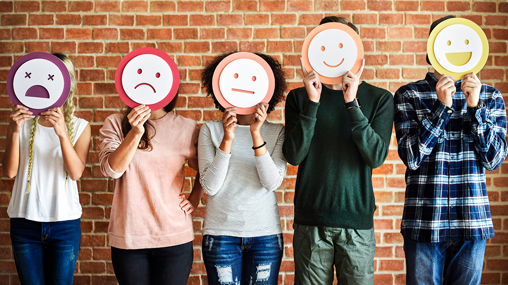 Image of five young people stood in front of a brick wall with each holding up a simplified mask face in front of their own face. The faces get progressively happier from left to right. 