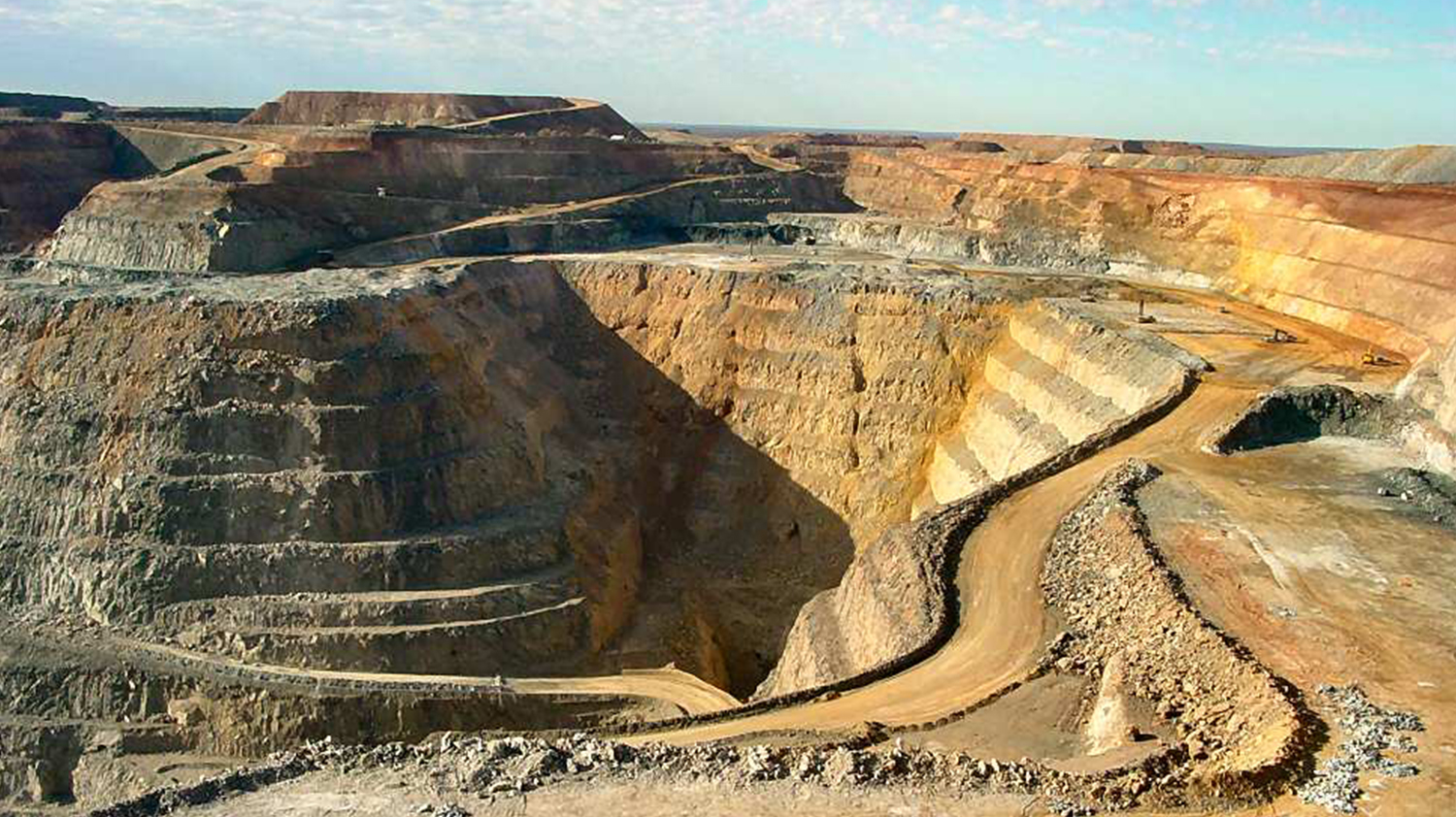 Photograph of an open pit mine in Australia where there is a deep, open pit in the centre. The surrounding landscape is yellow and golden in colour and the sky on the horizon is very pale blue.