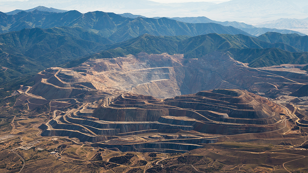 The image shows a view of the Bingham Canyon Mine diagonally from above. The immediate landscape is a rusty brown and gold colour and the mountains in the background have dark green fauna on them. The landscape has lots of curves where the mine is, winding around overlapping each other.