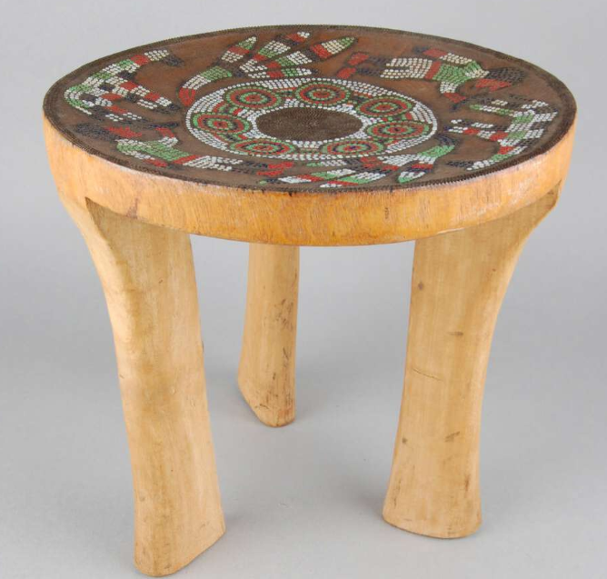 Three-legged carved wooden stool with circular seat, darkened and inlaid with human, animal and plant motifs in red, green, black and white glass beads and outlined with small metal staples.