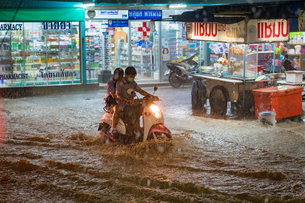 Two people on a motor scooter riding through a flooded urban street at night with illuminated shopfronts