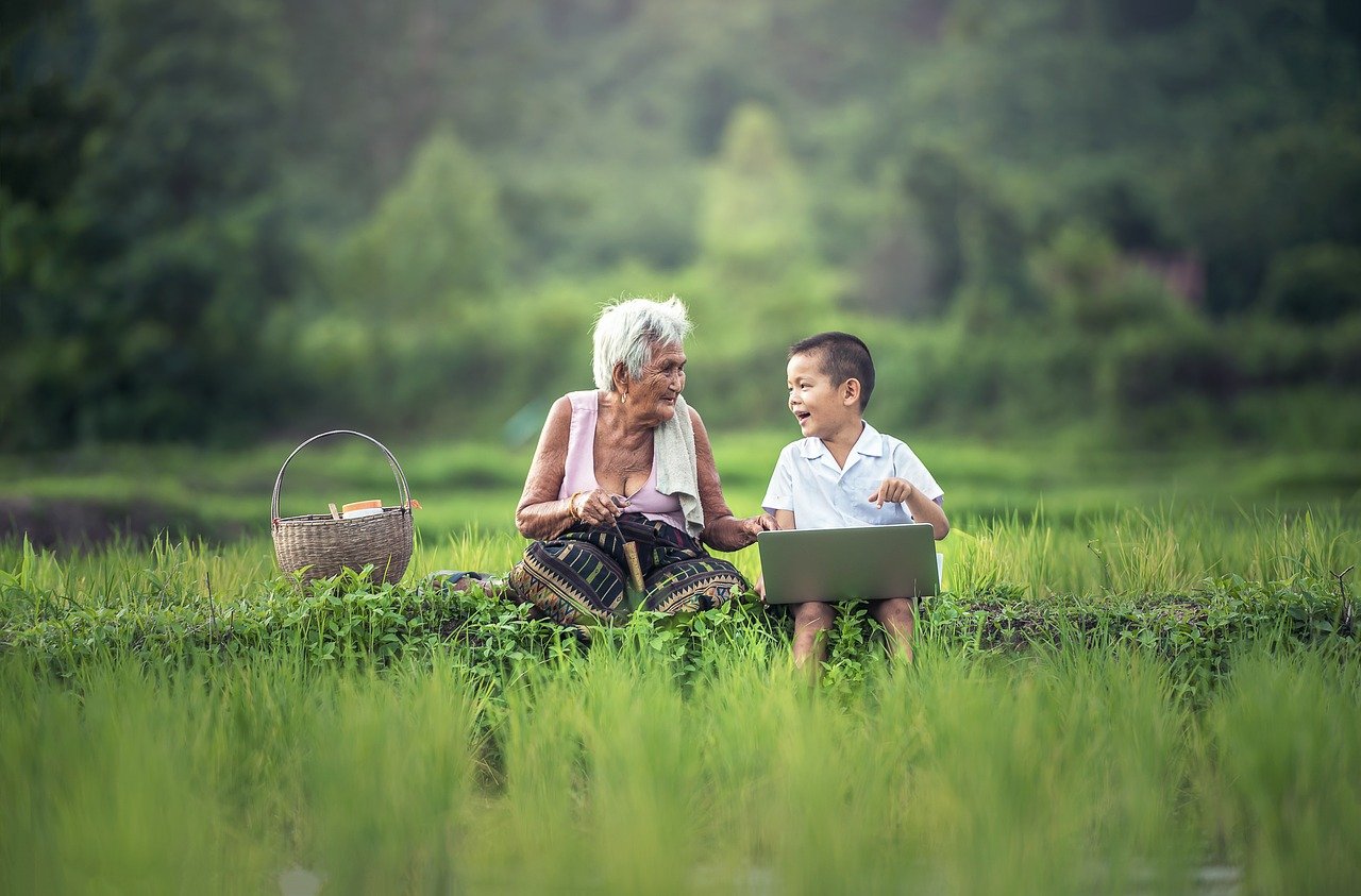 An old woman sits with a laughing child on their laptop, surrounded by rural nature