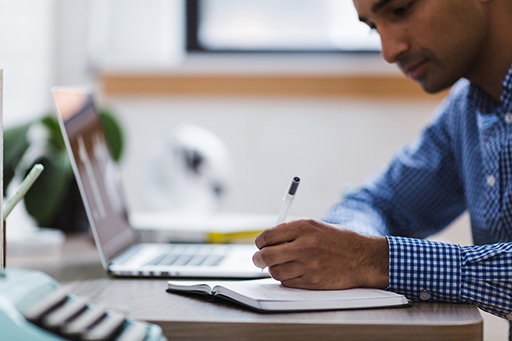 Photo of a person writing on a notebook with a laptop next to them.