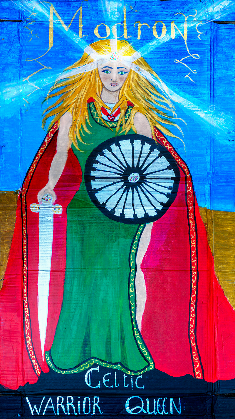 A painting of the mother goddess Modron depicted as a female warrior holding a sword and shield. Modron is wearing a bright green dress and red cloak, she has long blonde hair and light is emitting from her head dress. Above her is the text ‘Modron’, while below her feet are the words ‘Celtic Warrior Queen’.