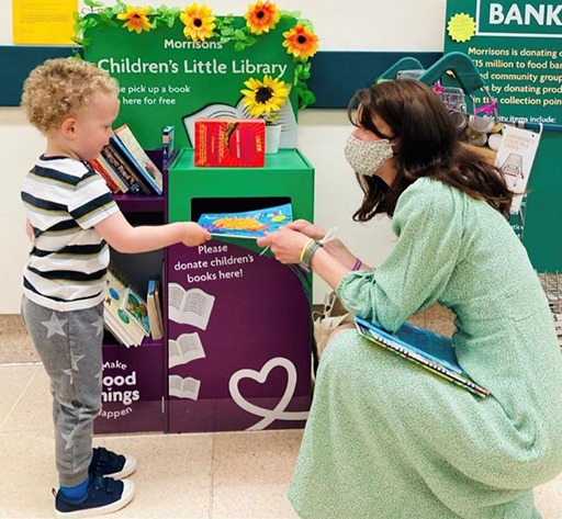 An adult kneeling down to help a child put a book in a library donation box.
