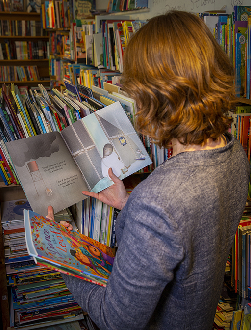 The back of an adult looking at a children’s book surrounded by stacked books.