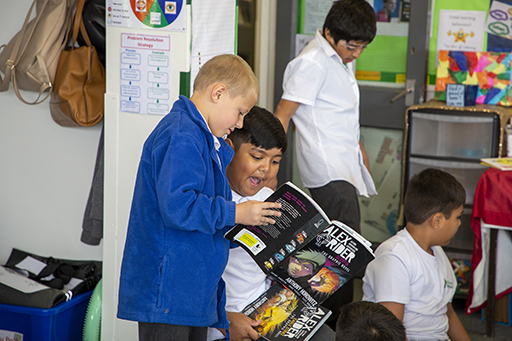 Photo of a classroom, focusing on 2 boys. 1 is showing something in an Alex Rider book to the other.