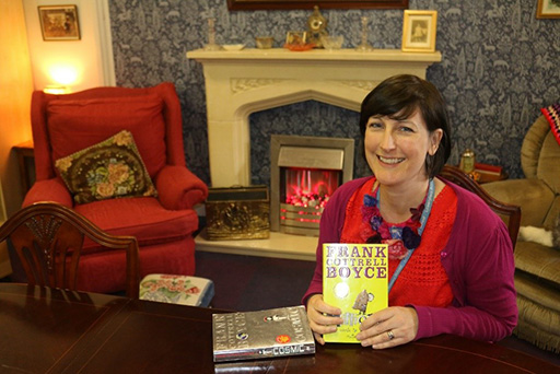 A woman smiling at the camera holding a book. Behind her is a fireplace and two armchairs.