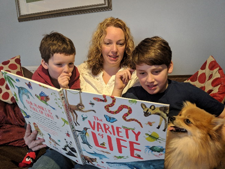 A parent with her two children reading the book ‘The variety of life’ on the sofa.