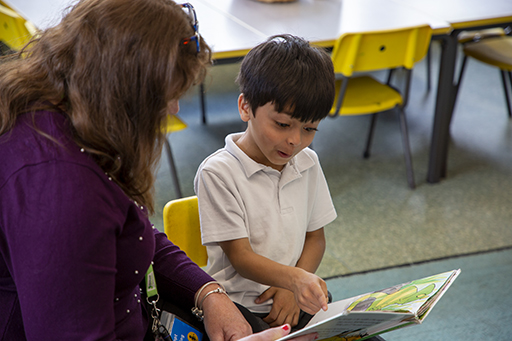 A young boy reading with an adult. He is pointing at the book and has an excited expression on his face.