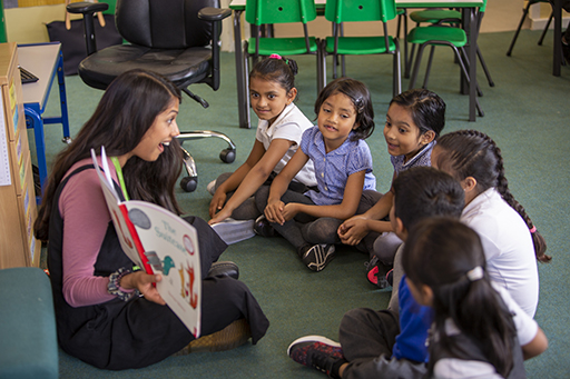 An adult sat on the classroom floor reading aloud to a group of children.