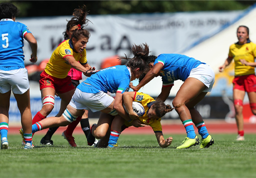 The image shows action from a rugby match. One player is being tackled by two players from the opposition. The player being tacked is falling head first towards the ground.