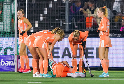 The image shows a group of female hockey players wearing orange strips. One of the hockey players is lying injured on the group while three other players lean over her.