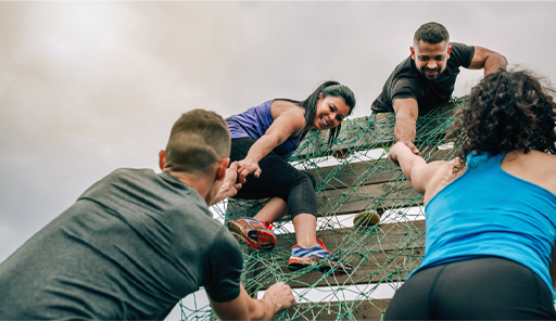 The image shows four people climbing a wooden obstacles. There are two people at the top of the obstacle who are reaching down with their hands to help the other two people up the obstacle. This image represents the value of team work.