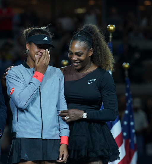 The image shows two tennis players, Serena Williams, and Naomi Osaka. They are having a private conversation and Naomi Osaka is laughing at something that Serena Williams is saying.