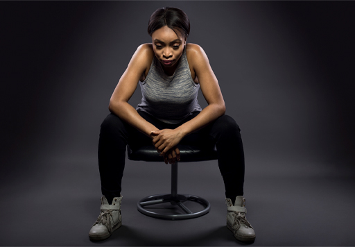The image shows a female in athletic clothing sitting on a stool. She is leaning forwards and has her hands crossed in front of her. She is deep in thought.