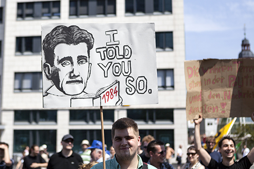 Person holding a banner at a demonstration with a drawing of George Orwell holding 1984. The banner reads ‘I told you so’